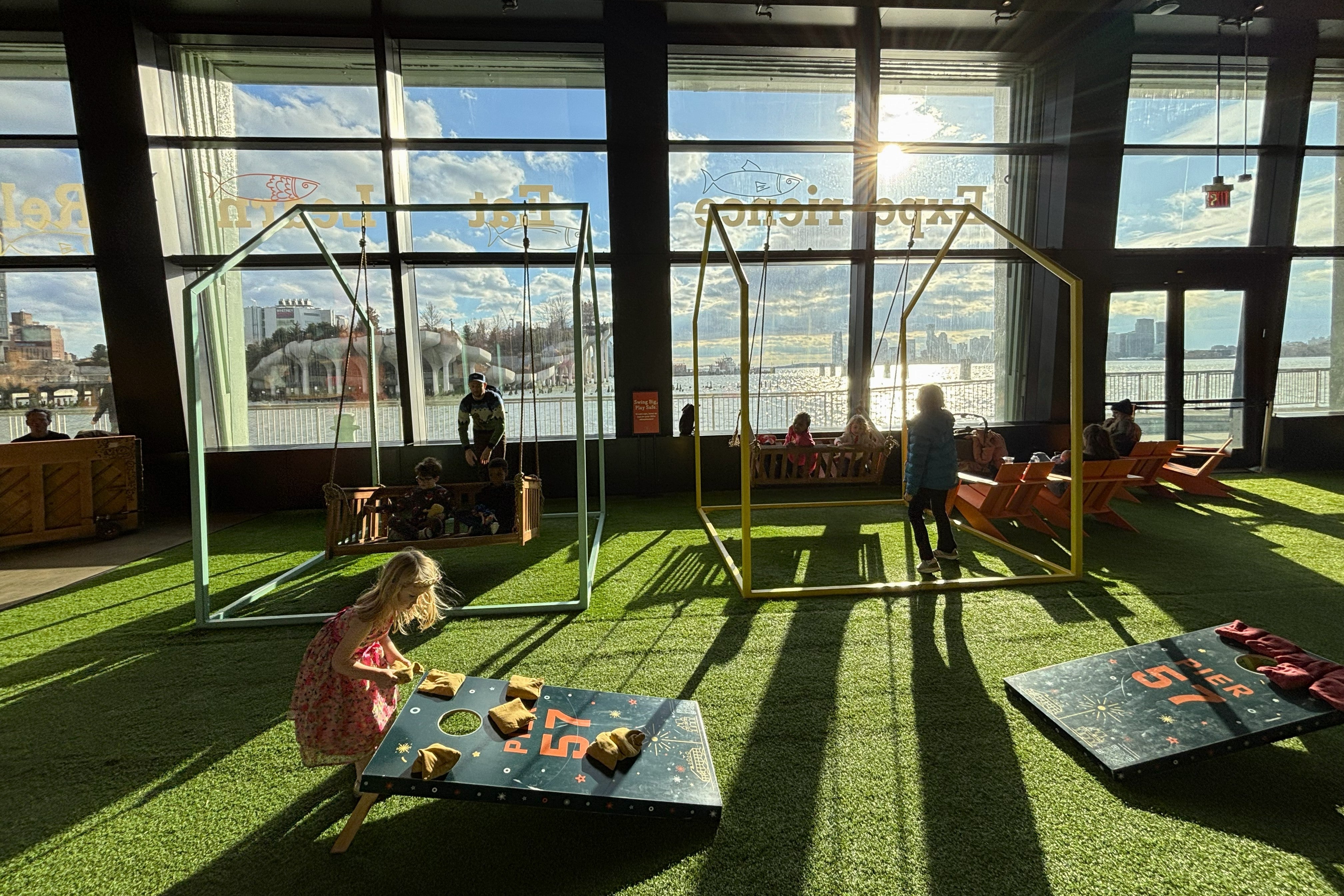 Indoor play spaces in Lower Manhattan: Indoor grassy playground with kids on swings and playing cornhole at Pier 57